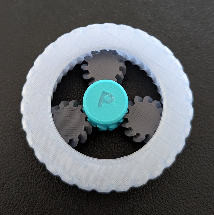 Photo of the printed Orbit Gearwheel, attempt 8, from above: a clear ring around three dark grey planet gears and a teal sun cap embossed with a P